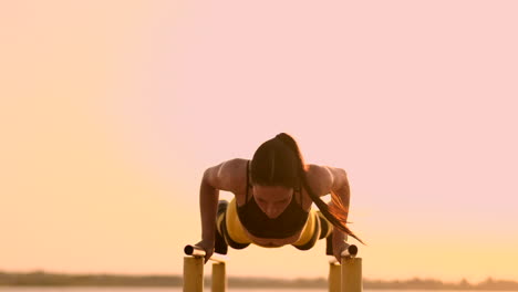 Beautiful-slender-woman-athlete-in-a-black-top-and-yellow-pants-at-sunset-performs-pushups-on-a-parallel-horizontal-bar-in-slow-motion.